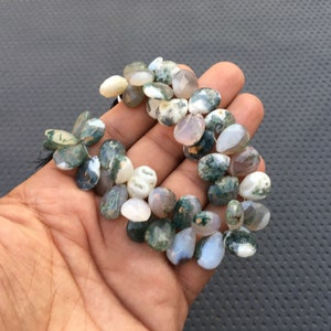 May include: A strand of green moss agate gemstone beads. The beads are a mix of light and dark green, white, and brown colors. The beads are teardrop shaped and have a natural, polished finish.