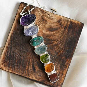 Puede incluir: Un collar de cadena de plata con siete piedras preciosas de diferentes colores. Las piedras preciosas están dispuestas en línea, comenzando con una amatista púrpura, luego una topacio azul, una esmeralda verde, una aguamarina azul claro, un peridoto verde, una citrina naranja y un granate rojo.