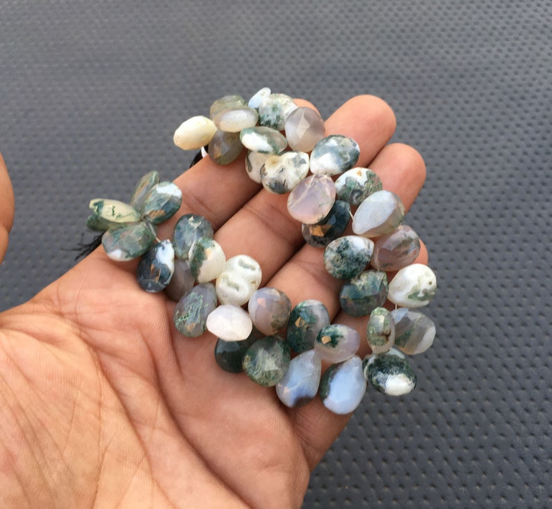 May include: A strand of green moss agate gemstone beads. The beads are a mix of light and dark green, white, and brown colors. The beads are teardrop shaped and have a natural, polished finish.