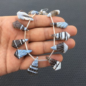 Puede incluir: Un hilo de cuentas de piedras preciosas talladas. Las cuentas son una mezcla de azul, blanco y marrón, con intrincados patrones geométricos y en forma de hoja. Las cuentas están ensartadas en alambre plateado, creando un componente de joyería único.