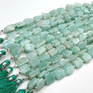 May include: A strand of light green aquamarine gemstone beads, each bead is a unique, irregular shape. The beads are strung on a white thread and are accented with silver tassels.