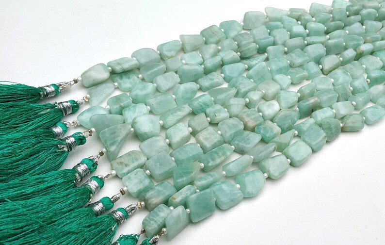 May include: A strand of light green aquamarine gemstone beads, each bead is a unique, irregular shape. The beads are strung on a white thread and are accented with silver tassels.