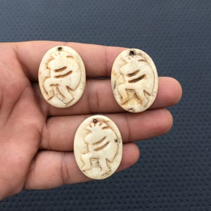 May include: Three oval-shaped pendants made of bone with a carved design of a stylized animal. The pendants are off-white with a slightly yellowed hue.