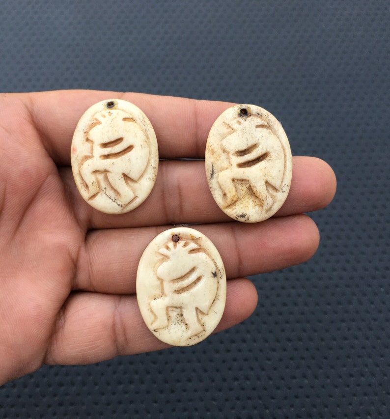 May include: Three oval-shaped pendants made of bone with a carved design of a stylized animal. The pendants are off-white with a slightly yellowed hue.