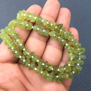 May include: A strand of green peridot gemstone beads with white spacer beads. The beads are faceted and have a rough, natural look.