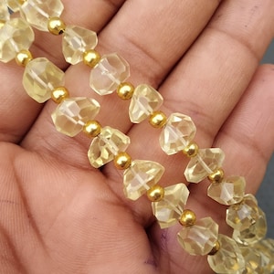 May include: Two strands of faceted, pale yellow gemstone beads with gold-coloured spacer beads. The beads are held in a hand, with a dark grey background.