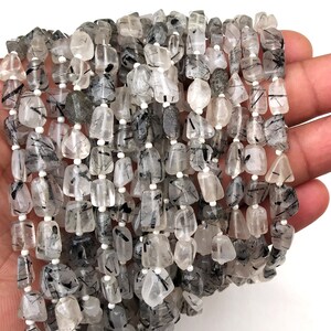 May include: A strand of black tourmaline beads with clear quartz inclusions. The beads are faceted and have a natural, rough texture. The beads are strung on a white thread.