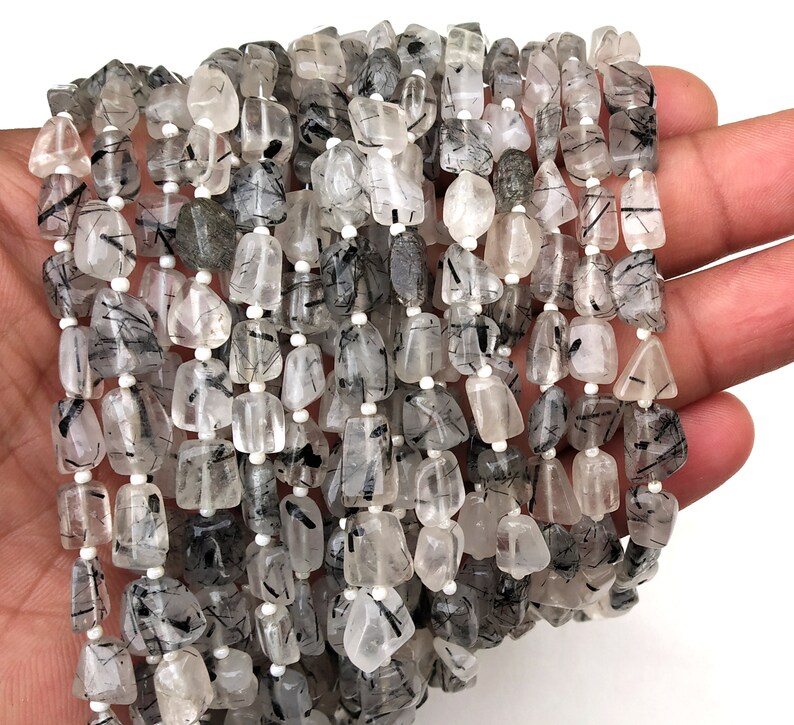 May include: A strand of black tourmaline beads with clear quartz inclusions. The beads are faceted and have a natural, rough texture. The beads are strung on a white thread.