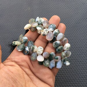 May include: A strand of green moss agate gemstone beads. The beads are a mix of light green, white, and gray colors. The beads are faceted and have a pear shape.