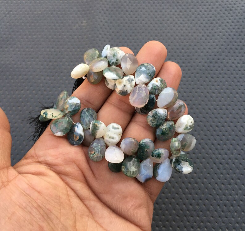 May include: A strand of green moss agate gemstone beads. The beads are a mix of light green, white, and gray colors. The beads are faceted and have a pear shape.