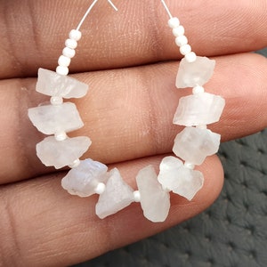 Puede incluir: Una hebra de cuentas de piedra lunar blanca con un alambre de plata y cuentas espaciadoras blancas. Las cuentas de piedra lunar son de forma irregular y tienen un color blanco lechoso.