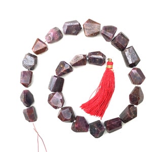 May include: A strand of faceted, dark red-brown gemstone beads. The beads are irregularly shaped with multiple facets, strung together with a red tassel at the end. The background is plain white, highlighting the beads' color and texture.