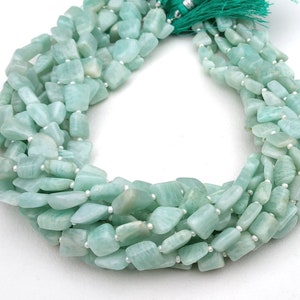 May include: A strand of light green gemstone beads, each bead is a slightly irregular, flat, rectangular shape. The beads are strung on a white thread.