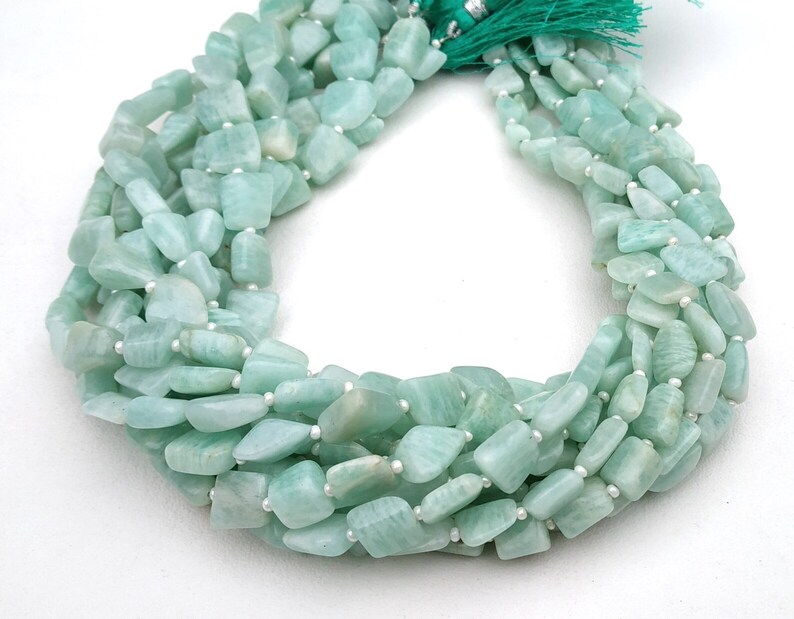 May include: A strand of light green gemstone beads, each bead is a slightly irregular, flat, rectangular shape. The beads are strung on a white thread.