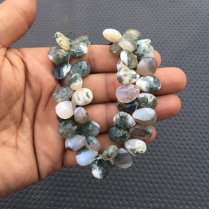 May include: A strand of green moss agate beads. The beads are a mix of light green, white, and gray with a mottled pattern. The beads are oval shaped and have a smooth, polished finish.