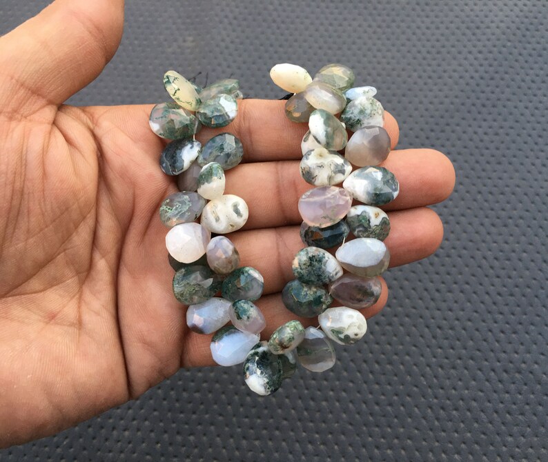 May include: A strand of green moss agate beads. The beads are a mix of light green, white, and gray with a mottled pattern. The beads are oval shaped and have a smooth, polished finish.