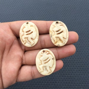 May include: Three oval-shaped pendants made of white bone with a carved design of a stylized animal figure. The pendants have a hole at the top for hanging.