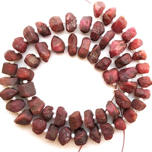 May include: A strand of red ruby gemstone beads, each bead is irregularly shaped and has a rough, natural finish. The beads are strung on a white thread with small white spacer beads.