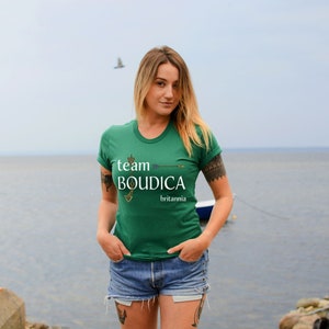 Pu&ograve; includere: Una donna indossa una maglietta verde con il testo "team BOUDICCA britannia" in lettere bianche. &Egrave; in piedi di fronte a uno specchio d'acqua.