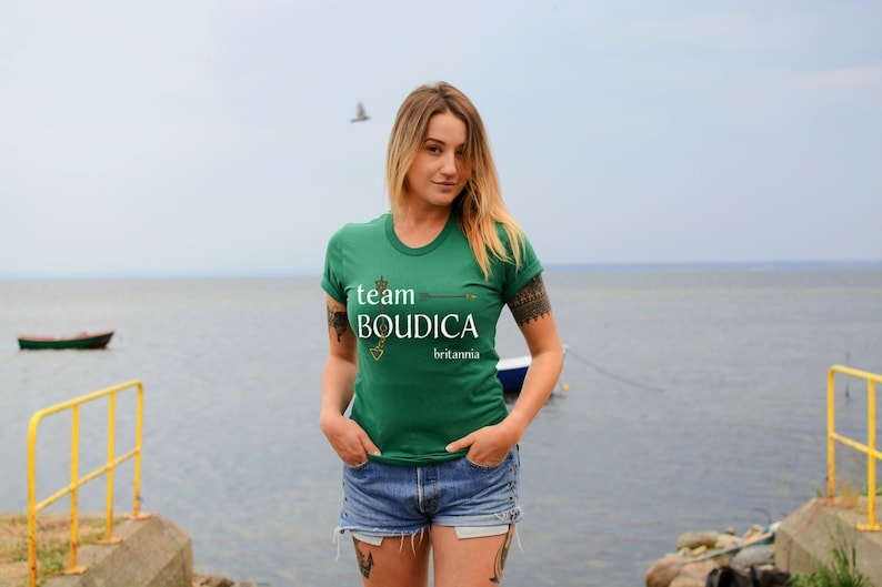 Pu&ograve; includere: Una donna indossa una maglietta verde con il testo "team BOUDICCA britannia" in lettere bianche. &Egrave; in piedi di fronte a uno specchio d'acqua.