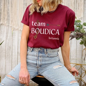 Pu&ograve; includere: Una maglietta color granata con il testo "team BOUDICCA britannia" e una grafica di freccia dorata.
