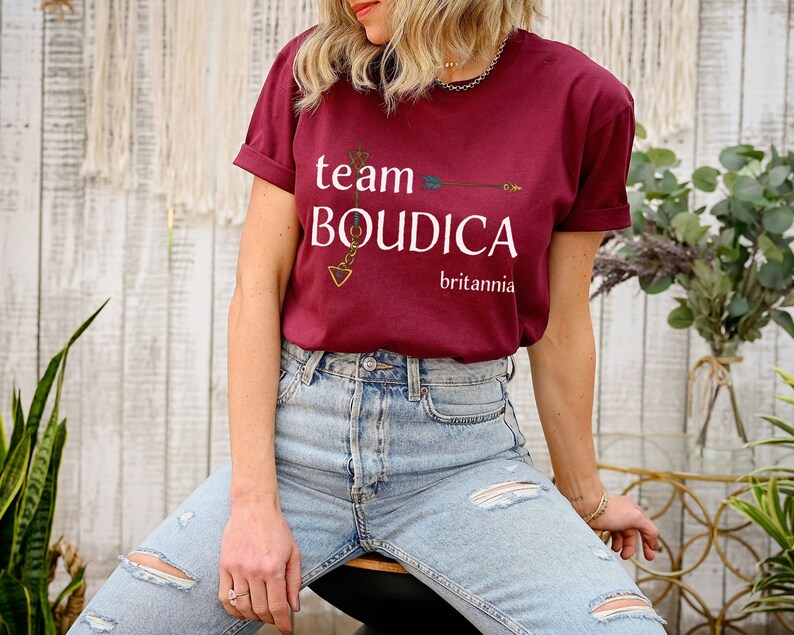 Pu&ograve; includere: Una maglietta color granata con il testo "team BOUDICCA britannia" e una grafica di freccia dorata.
