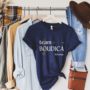 Pu&ograve; includere: Una maglietta blu navy con il testo "team BOUDICA britannia" e una grafica di freccia dorata.