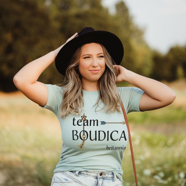 Pu&ograve; includere: Una donna indossa una maglietta blu chiaro con il testo "team BOUDICA britannia" e un design a chiave dorata. Indossa un cappello nero e ha lunghi capelli biondi.