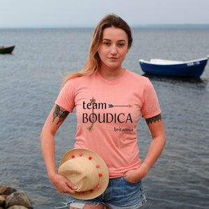 Pu&ograve; includere: Una donna indossa una maglietta color pesca con il testo "team BOUDICA britannia" e una grafica a freccia. Tiene in mano un cappello di paglia con pompon rosa.