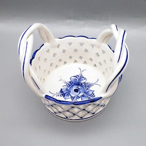 May include: A white ceramic basket featuring a blue hand-painted floral design at the center. The bowl has a lattice-style openwork pattern on the sides and two twisted handles with blue stripes. The rim is scalloped with a blue painted edge.