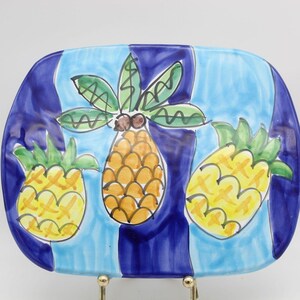 May include: A rectangular ceramic platter featuring a hand-painted design of three pineapples with green leaves and yellow and orange bodies. The background consists of alternating vertical stripes in light blue and deep navy blue paint.
