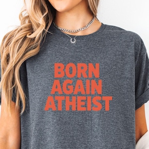 May include: Dark gray t-shirt with the words "BORN AGAIN ATHEIST" in orange block letters. The shirt has short sleeves and a crew neck. The model is wearing a white jacket over the shirt.