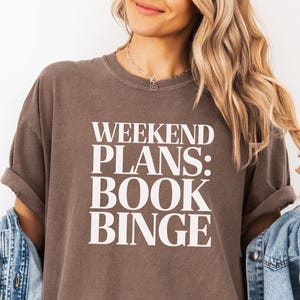 May include: Espresso-colored t-shirt with the text "WEEKEND PLANS: BOOK BINGE" in white. The shirt is worn with a light blue denim jacket. The shirt is made of Comfort Colors material.