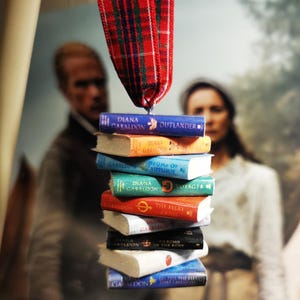 May include: A stack of miniature books with colourful covers and titles such as "Outlander" and "Voyager" hangs from a red and green plaid ribbon. The books are stacked vertically, with a blurred background.