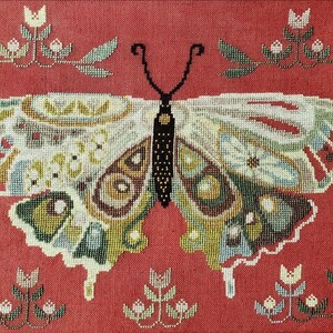 May include: A cross-stitch embroidery on a red fabric background featuring a large central butterfly with intricate patterns in shades of white, green, blue, and brown. The design is surrounded by small, symmetrical floral motifs in the corners.