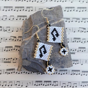 Peut inclure: Boucles d'oreilles perlées faites à la main avec un motif géométrique. Les boucles d'oreilles présentent des panneaux carrés perlés blancs et noirs avec des accents dorés et une petite fleur perlée pendante. Les boucles d'oreilles sont exposées sur une pierre grise.