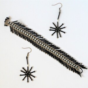 May include: A jewellery set featuring a bracelet and two pairs of earrings. The bracelet and earrings are black with silver accents. The earrings have a starburst design. The jewellery is displayed on a white surface.