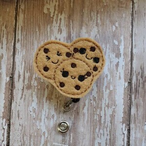 May include: A brown felt cookie charm with a smiling face and sunglasses. The charm is attached to a clear plastic retractable badge holder.