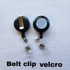 May include: Two black retractable badge holders with a belt clip and a velcro backing. The holders are on a white brick background.