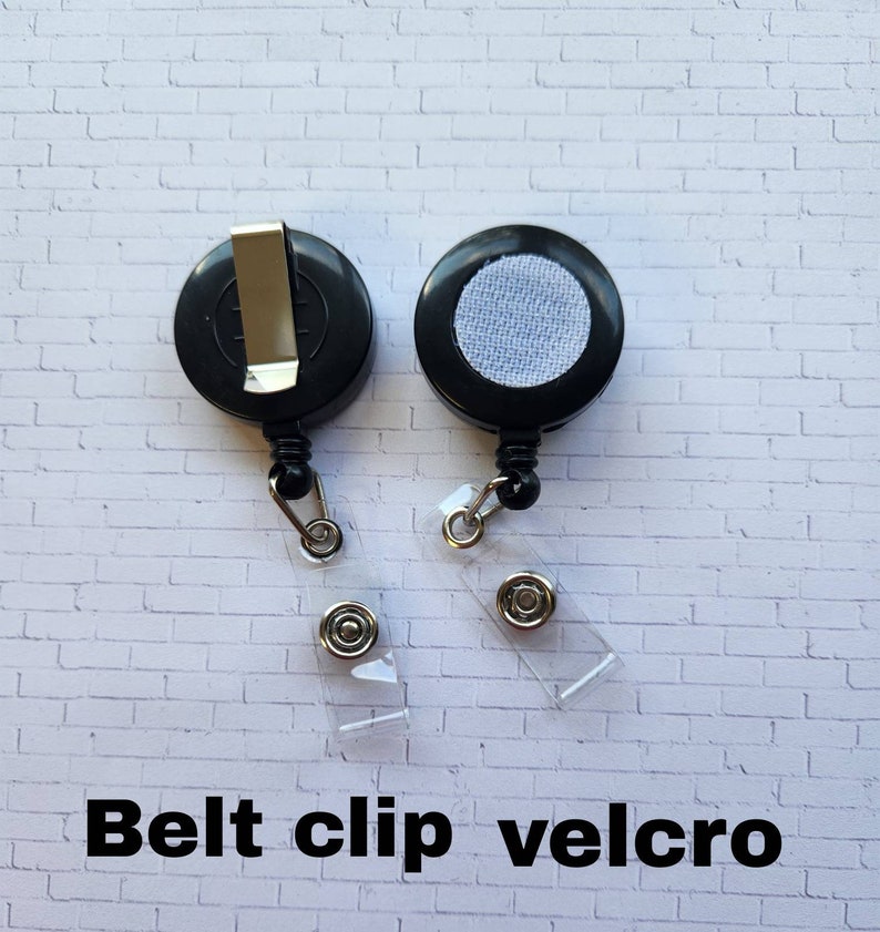 May include: Two black retractable badge holders with a belt clip and a velcro backing. The holders are on a white brick background.