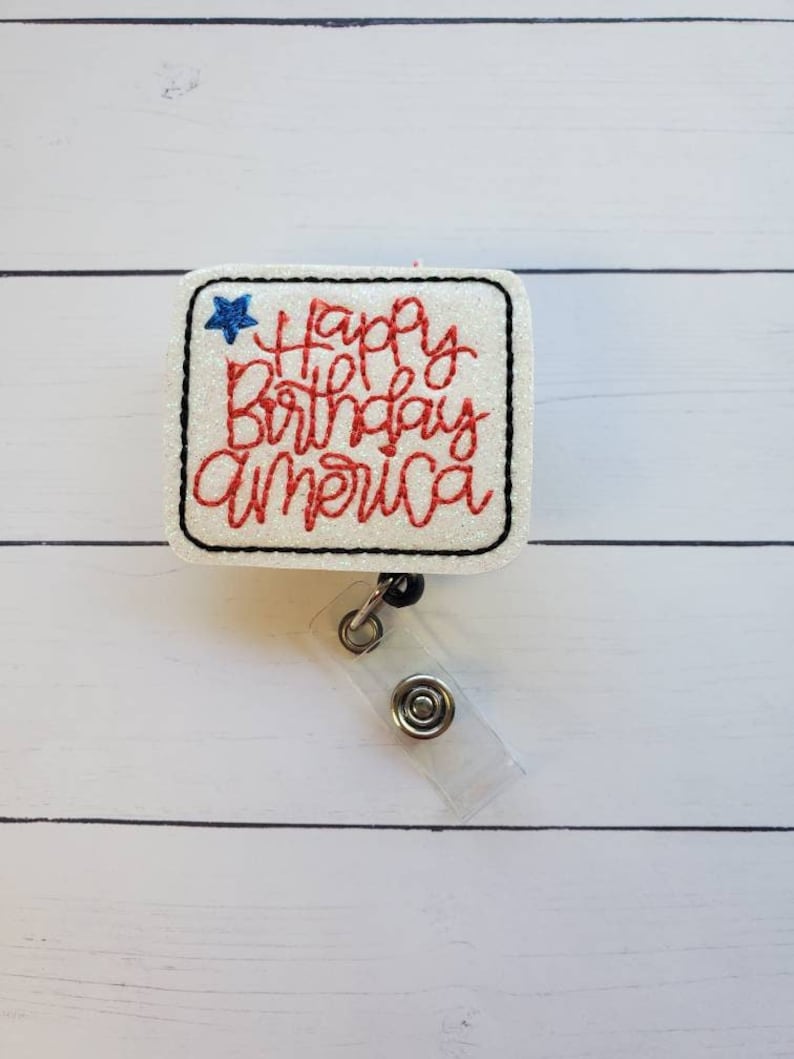 K&ouml;nnte beinhalten: Wei&szlig;er Glitzer-Badgehalter mit einem roten und blauen gestickten Design, auf dem "Happy Birthday America" und ein blauer Stern stehen. Der Badgehalter ist an einem durchsichtigen, einziehbaren Badge-Band befestigt.