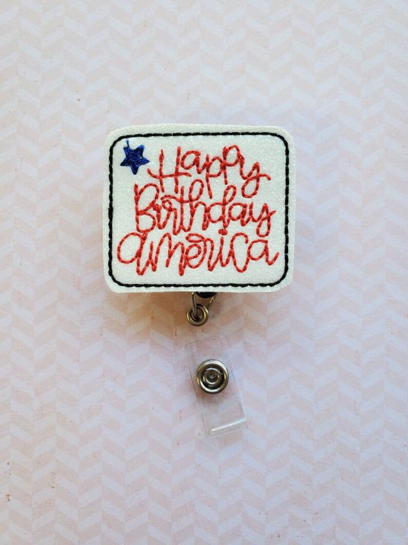 K&ouml;nnte beinhalten: Wei&szlig;es quadratisches Abzeichen mit einem blauen Stern und rotem gesticktem Text mit der Aufschrift "Happy Birthday America". Das Abzeichen ist an einem durchsichtigen, einziehbaren Ausweishalter aus Kunststoff mit einem silbernen Clip befestigt.