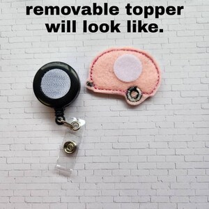 May include: A close-up of a pink felt camper van with a white circle in the middle. The camper van is attached to a black retractable badge reel with a clear plastic strap. The text "This is what a removable topper will look like." is visible at the top of the image.