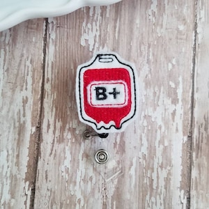 May include: White embroidered badge with a red blood bag design and the text "B+" inside. The badge is attached to a retractable badge holder.