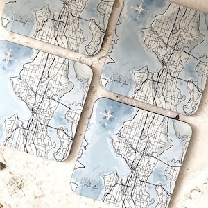 May include: Set of four blue and white coasters featuring a map of Seattle, Washington. Each coaster has a compass rose and the text "Seattle, Washington" in a handwritten font.
