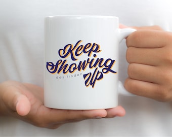 May include: White ceramic mug with a blue and yellow design that says "Keep Showing Up" with the text "des linden" below.