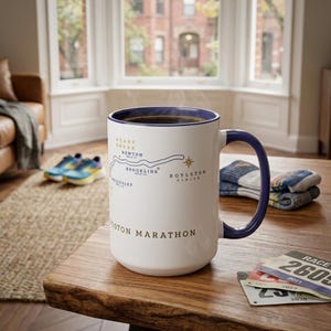 May include: A white ceramic mug with a blue rim and handle, filled with dark liquid. The mug features a map of the Boston Marathon route with text. Running shoes and race bibs are also visible.