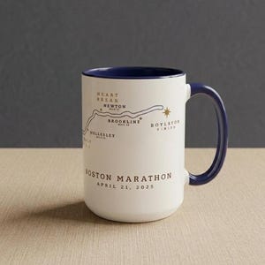 May include: White ceramic mug with a navy blue handle and interior rim. The mug features a map of the Boston Marathon route, with key locations labeled. Text on the mug reads "Boston Marathon April 21, 2025."