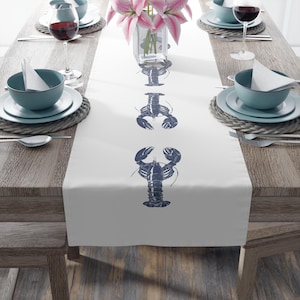 Puede incluir: Camino de mesa blanco con tres ilustraciones de langostas azul marino. El camino de mesa está colocado sobre una mesa de madera preparada para una comida, con platos, cuencos, cubiertos y copas de vino. Un jarrón con flores rosas está en el centro.