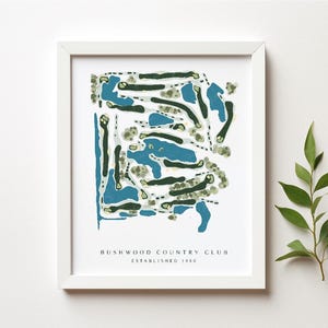 May include: White framed print of a golf course map with blue and green details. The text "Bushwood Country Club Established 1980" is printed at the bottom. A sprig of green leaves is placed to the right of the frame.